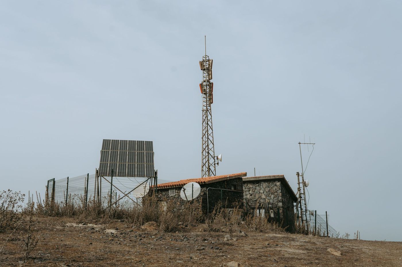 An isolated telecom site on a dry ridge: lattice tower, sector antennas, solar panel and a small equipment shelter.