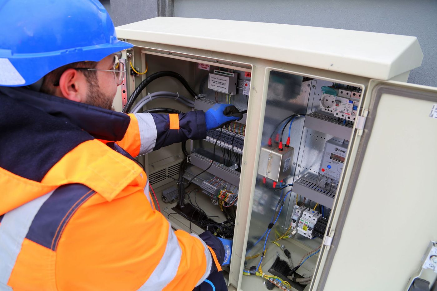 A technician in a high-visibility jacket and helmet working inside an open outdoor equipment cabinet.